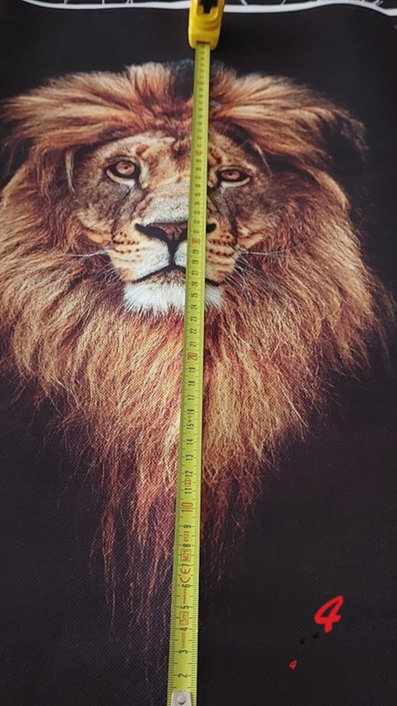 May include: A black fabric panel featuring a detailed, realistic portrait of a lion with a full, golden-brown mane. A yellow measuring tape is placed vertically over the centre of the lion's face, showing a length of approximately 32 centimetres.