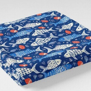May include: Folded fabric with a repeating pattern of white and blue fish, red seashells, and aquatic elements on a dark blue background. The fabric is stacked, showing the layered design. The overall theme is nautical.