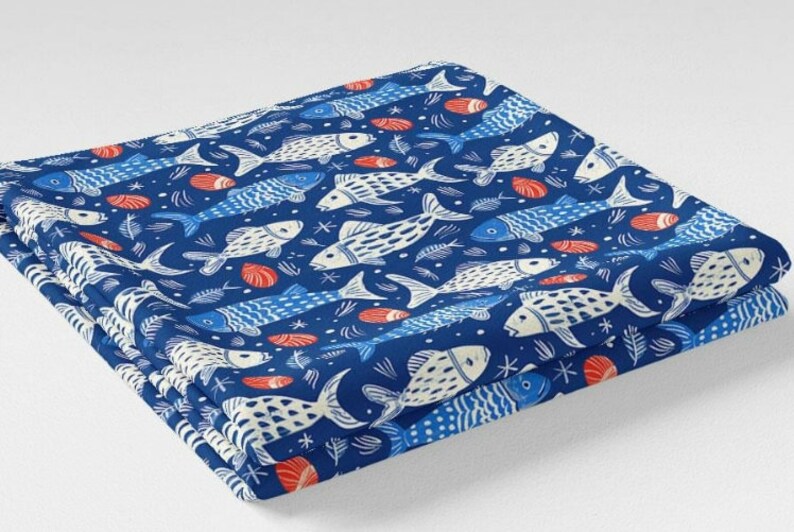May include: Folded fabric with a repeating pattern of white and blue fish, red seashells, and aquatic elements on a dark blue background. The fabric is stacked, showing the layered design. The overall theme is nautical.