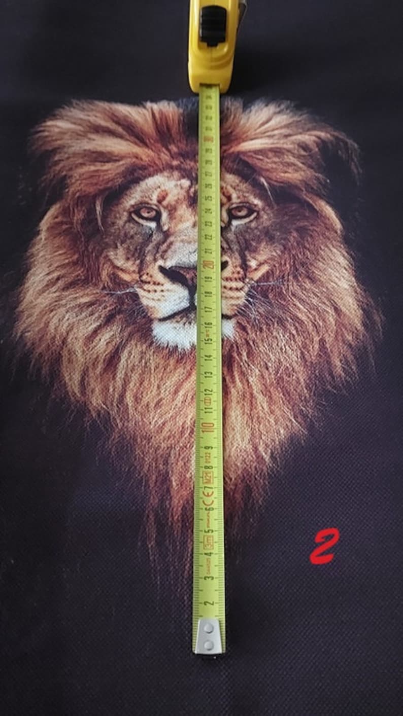 May include: A black fabric featuring a detailed, realistic print of a lion's face with a full, brown mane. A yellow measuring tape is positioned vertically over the centre of the lion's face, showing a length of approximately 30 cm.