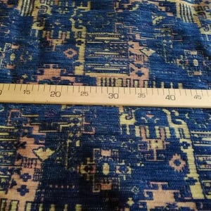 May include: A blue fabric featuring a woven geometric pattern in shades of tan and light pink. A wooden ruler is placed horizontally across the textile, showing measurements from 15 to 45 centimetres, indicating the scale of the intricate design.
