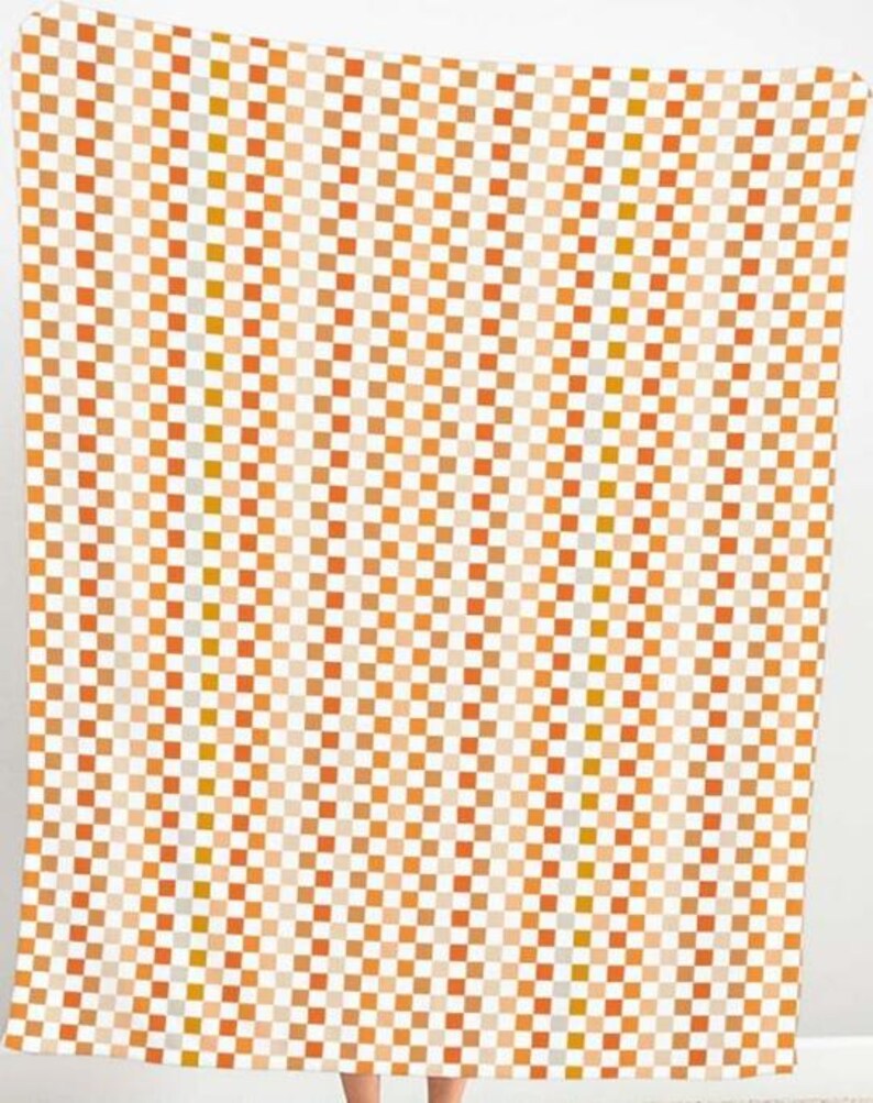 K&ouml;nnte beinhalten: Eine Decke mit einem sich wiederholenden Schachbrettmuster in Orange-, Beige- und Wei&szlig;t&ouml;nen. Die Quadrate sind gleichm&auml;&szlig;ig gro&szlig; und erzeugen ein ansprechendes Design. Die Decke besteht aus einem weichen Material und dient wahrscheinlich der W&auml;rme und dem Komfort.