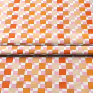 K&ouml;nnte beinhalten: Nahaufnahme eines Stoffes mit sich wiederholendem Schachbrettmuster. Die Quadrate wechseln zwischen Orange-, Rosa- und Beiget&ouml;nen, mit eingestreuten wei&szlig;en Quadraten. Der Stoff ist unten gefaltet und zeigt die Kontinuit&auml;t des Musters.