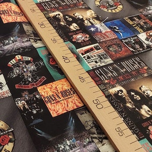 May include: Close-up of fabric printed with Guns N' Roses imagery, including album covers and band photos. The design incorporates the band's logo, roses, and text like "Appetite for Destruction". A wooden ruler, marked in centimetres, lies across the fabric.