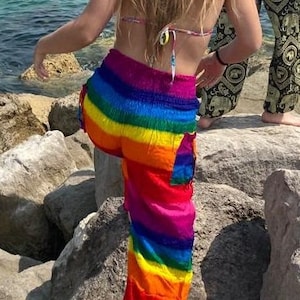 Pu&ograve; includere: Pantaloni a righe arcobaleno con colori vivaci: rosso, arancione, giallo, verde, blu e viola. I pantaloni larghi hanno una tasca laterale. La foto mostra i pantaloni all'aperto, vicino al mare.
