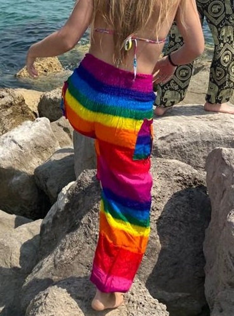 Pu&ograve; includere: Pantaloni a righe arcobaleno con colori vivaci: rosso, arancione, giallo, verde, blu e viola. I pantaloni larghi hanno una tasca laterale. La foto mostra i pantaloni all'aperto, vicino al mare.