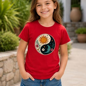 May include: A red t-shirt with a Yin and Yang design featuring a sun, moon, stars, and floral elements. The shirt is worn with blue denim shorts. The design is in the centre of the t-shirt.
