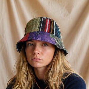 Puede incluir: Un sombrero de pescador confeccionado con varios parches de tela que presentan una mezcla de texturas y patrones. El diseño incluye secciones de encaje verde, tela negra con rayas verticales rojas y blancas y un panel con estampado floral morado.