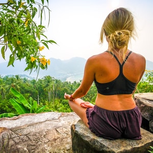 Pu&ograve; includere: Una persona con un reggiseno sportivo nero e pantaloncini viola &egrave; seduta in posizione meditativa su una grande roccia, con vista su un paesaggio tropicale. La scena comprende lussureggianti alberi verdi, una baia e montagne in lontananza. Fiori gialli e fogliame verde incorniciano la vista.