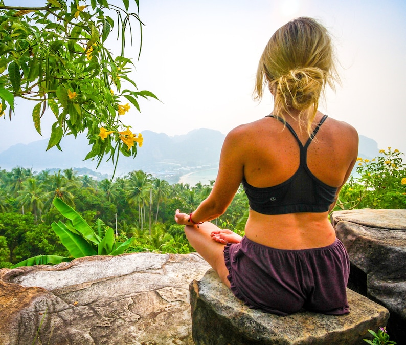 Pu&ograve; includere: Una persona con un reggiseno sportivo nero e pantaloncini viola &egrave; seduta in posizione meditativa su una grande roccia, con vista su un paesaggio tropicale. La scena comprende lussureggianti alberi verdi, una baia e montagne in lontananza. Fiori gialli e fogliame verde incorniciano la vista.