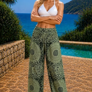 May include: Green and black patterned harem trousers with a wide leg. The trousers feature a detailed circular design. The model is wearing a white halter top and brown sandals. The background includes a pool, sea, and trees.