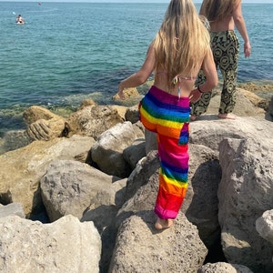 Pu&ograve; includere: Una persona con pantaloni a righe arcobaleno e un top bikini &egrave; in piedi su rocce vicino al mare. Un'altra persona con pantaloni fantasia cammina nelle vicinanze. La scena &egrave; ambientata in una giornata di sole con cielo blu chiaro e acqua.