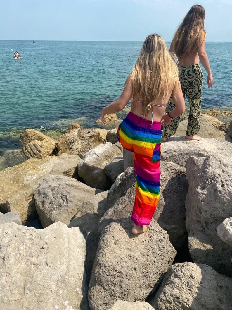 Pu&ograve; includere: Una persona con pantaloni a righe arcobaleno e un top bikini &egrave; in piedi su rocce vicino al mare. Un'altra persona con pantaloni fantasia cammina nelle vicinanze. La scena &egrave; ambientata in una giornata di sole con cielo blu chiaro e acqua.