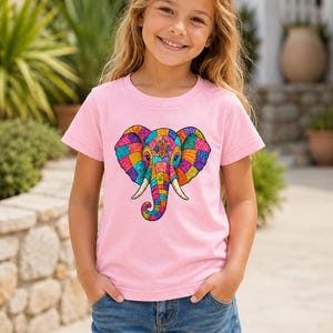 May include: A light pink t-shirt with a vibrant elephant design. The elephant is composed of colourful geometric patterns in blue, green, yellow, orange, and pink. The shirt is worn by a person with blonde hair and blue denim shorts.