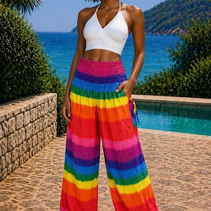 Könnte beinhalten: Eine Person trägt ein weißes Neckholder-Top und eine Regenbogen-Haremshose. Die Hose hat horizontale Streifen in Rot, Orange, Gelb, Grün, Blau und Lila. Der Hintergrund umfasst einen Pool, das Meer und einen blauen Himmel.