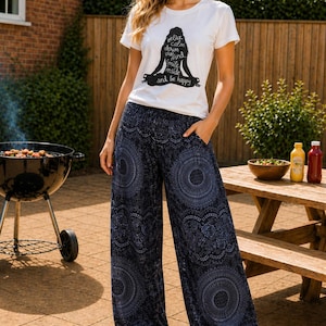 May include: A woman wearing a white t-shirt with a black graphic of a meditating figure and the words "Relax, Calm, Deep, Breathe, Mind, Smile, Inside, and Be Happy." She is also wearing navy blue patterned pants and sandals.
