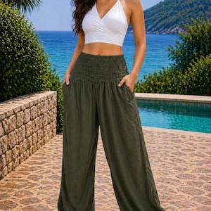 May include: A woman in a white halter top and olive green harem trousers. The trousers have a gathered waistband and pockets. She is standing on a stone patio with a pool and sea in the background. The outfit is casual and suitable for summer.