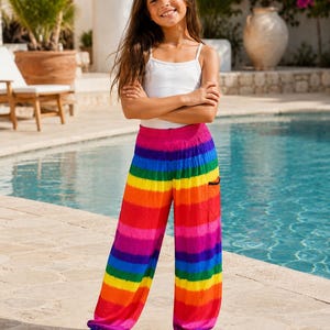 May include: A person wearing a white vest top and rainbow-striped harem trousers. The trousers have horizontal bands of red, orange, yellow, green, blue, and purple. The trousers have a relaxed fit and are cuffed at the ankles.