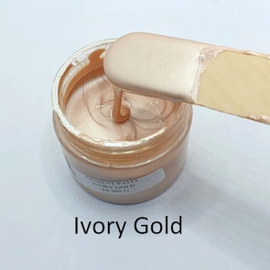 Peut inclure: Un petit pot de pâte pigmentée or ivoire. Le pot est ouvert et une spatule prélève de la pâte. L'étiquette sur le pot indique "Pigment Paste Ivory Gold PP-001-G".