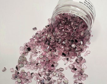以下が含まれることがあります： 「Crushed Glass Rose Pink 1/2-1/4」とラベル付けされた、砕いたガラスが入った瓶が白い表面にこぼれている。ガラスは薄いピンク色で、銀色の虹色の光沢がある。