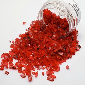 Op de afbeelding: Een glazen pot gevuld met rood gebroken glas. De pot is omgevallen en het glas valt op een wit oppervlak. Het etiket op de pot staat "Mirrored Glass Crushed Red  MIC-0027".