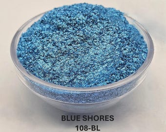 May include: A clear glass bowl filled with shimmering blue powder. The powder has a textured appearance and reflects light, creating a sparkling effect. The text "BLUE SHORES" and "108-BL" are printed at the bottom of the image.