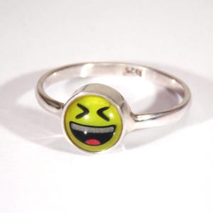 May include: A silver ring featuring a yellow emoji face with a wide open mouth and laughing eyes. The ring has a polished finish and a simple band. The emoji face is centered on the ring and is a bright, cheerful design.