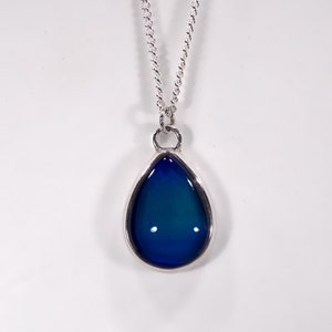 Puede incluir: Un collar de cadena de plata con un colgante en forma de lágrima. El colgante está hecho de vidrio azul que cambia de color en diferentes luces.
