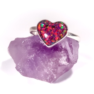 May include: A silver ring featuring a heart-shaped red opal gemstone. The ring is resting on a purple amethyst crystal.