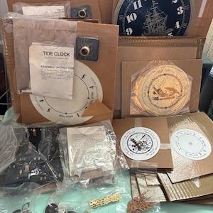 May include: A collection of tide clock components, including a navy blue clock face with a ship illustration, assembly instructions, and various hardware pieces. The clock face measures approximately 10 inches in diameter.