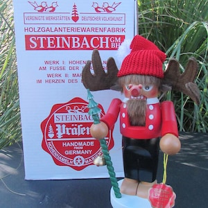 May include: A Steinbach nutcracker figurine in a red jacket, black pants, and a red knit cap, holding a green staff. The figurine has brown antlers and a white beard. A box with German text is in the background.