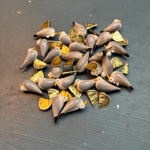 May include: A collection of small, gray and white ceramic bird figurines with black beaks and feet. Each bird is accompanied by a gold-colored label with text. The figurines are scattered on a blue-gray surface.