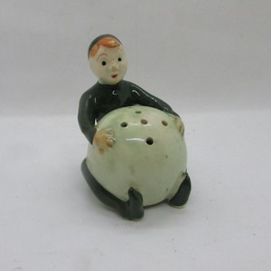 Puede incluir: Un salero de cerámica vintage con forma de figura sentada que sostiene una bola redonda de color verde claro con varios agujeros. La figura tiene una cara pintada, pelo naranja y viste ropa verde oscuro.