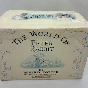 以下が含まれることがあります： ピーターラビットの世界、ビアトリクス・ポターによるヴィンテージボックスセット。クリーム色の箱にはウサギのイラストと黒と青の文字が描かれています。F. Warne & Co.によるオリジナルで認可された版です。