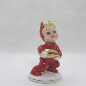 May include: A vintage ceramic figurine of a child in a red jumpsuit with gold accents, holding a gold football. The figurine has a white base with a gold trim and is set against a white background.