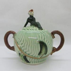 May include: A ceramic sugar bowl with a lid featuring a seated figure. The bowl is light green with brown handles and decorative accents. The lid has a textured design, and the figure is dressed in black.