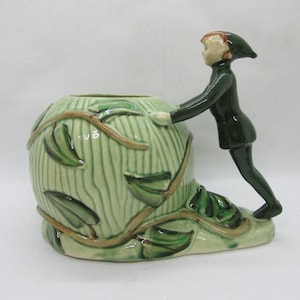 Puede incluir: Una maceta de cerámica con un diseño verde y marrón, que presenta la figura de un niño con un atuendo verde. La maceta tiene forma redonda con líneas verticales y detalles de hojas. La figura del niño está apoyada en la maceta.