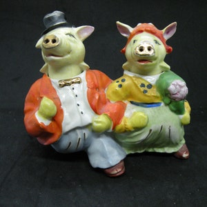 Puede incluir: Un par de figuras de cerdos de cerámica, uno con un sombrero de copa y una chaqueta roja, el otro con un vestido verde y una camisa amarilla con lunares negros.