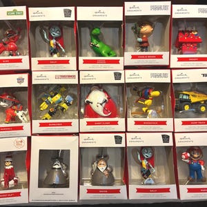 May include: A collection of Hallmark Christmas tree ornaments in boxes. The ornaments feature characters from Sesame Street, Peanuts, The Nightmare Before Christmas, Transformers, and Super Mario. Characters include Elmo, Sally, Charlie Brown, Snoopy, and Mario.