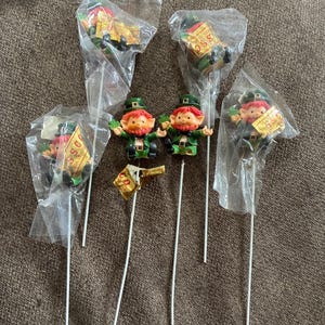 May include: Five leprechaun figurines attached to thin white sticks, each wearing a green hat, green jacket, and black belt. Two figurines are unwrapped, while three remain in clear plastic bags with gold labels reading SSC.