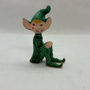Könnte beinhalten: Eine Keramikfigur eines sitzenden Elfen. Der Elf trägt ein grünes Outfit mit passendem spitzem Hut. Er hat große, spitze Ohren, rosige Wangen und blondes Haar. Die Figur ist ein dekoratives Objekt.