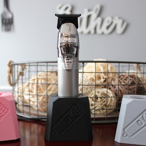 Puede incluir: Un cortapelos eléctrico plateado con un soporte negro. El soporte tiene un gráfico de un cortapelos en él. El cortapelos está sentado en una mesa de madera.