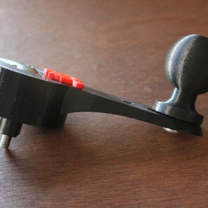 Op de afbeelding: Een zwarte 3D-geprinte camerabevestiging met een kogelkop en een rode knop. De bevestiging heeft een schroefgat om aan een statief te bevestigen.
