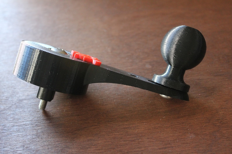 Op de afbeelding: Een zwarte 3D-geprinte camerabevestiging met een kogelkop en een rode knop. De bevestiging heeft een schroefgat om aan een statief te bevestigen.