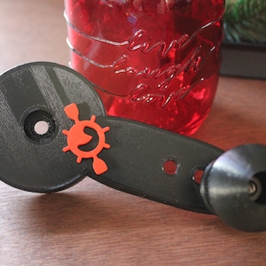 Op de afbeelding: Een zwarte 3D-geprinte versnellingspookknop met een rood en oranje stuurwiellogo. De knop heeft een ronde basis en een lange handgreep met een kleine zwarte knop aan het einde.