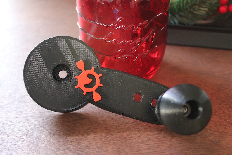 Op de afbeelding: Een zwarte 3D-geprinte versnellingspookknop met een rood en oranje stuurwiellogo. De knop heeft een ronde basis en een lange handgreep met een kleine zwarte knop aan het einde.