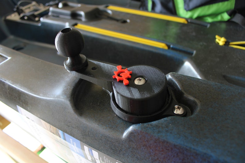 Op de afbeelding: Een zwart plastic kajakaccessoire met een rode knop in de vorm van een tandwiel. Het accessoire is gemonteerd op een zwarte kajak met een gele streep.