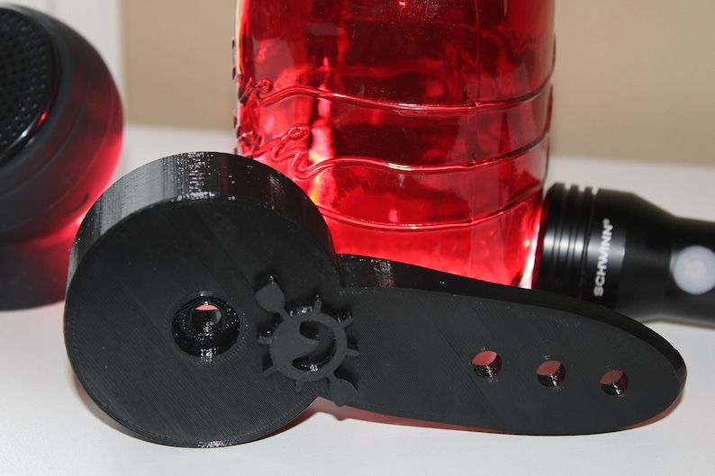 Op de afbeelding: Een zwart 3D-geprint onderdeel met een cirkelvormig ontwerp en drie gaten. Het onderdeel is gemaakt van plastic en heeft een gladde afwerking.