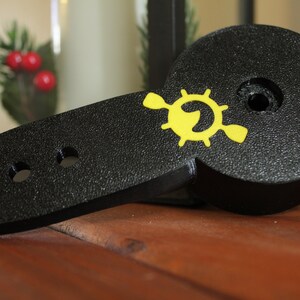 Op de afbeelding: Zwart plastic peddel met een geel logo van een stuurwiel met een vis erin. Het peddel heeft vier gaten aan de zijkant.