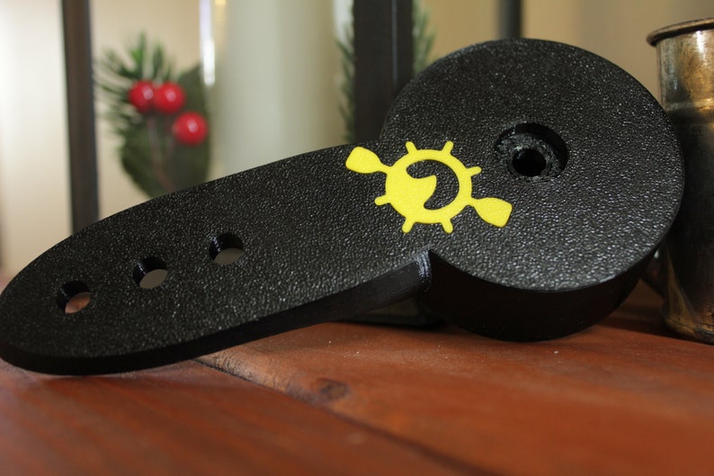 Op de afbeelding: Zwart plastic peddel met een geel logo van een stuurwiel met een vis erin. Het peddel heeft vier gaten aan de zijkant.